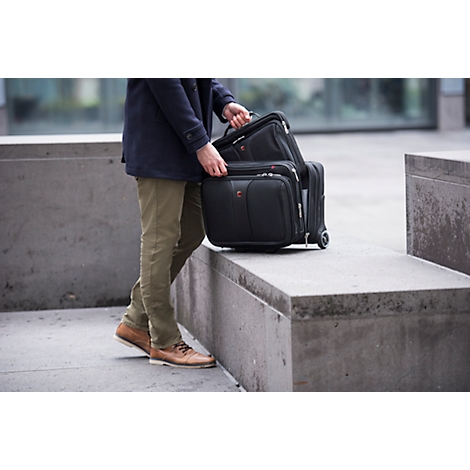 A person in olive pants and brown shoes carries several pieces of black luggage. The background is blurred.