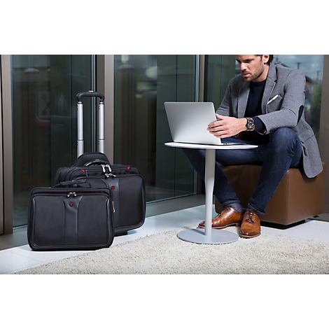 Man sits at a small table with a laptop. Suitcases are next to him. Glass wall in the background.