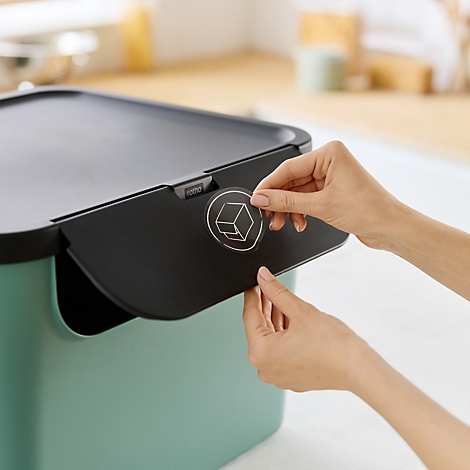 Nahaufnahme einer Person, die einen schwarzen Deckel eines türkisfarbenen Behälters öffnet, mit einem runden Logo mit einem Würfel darauf.