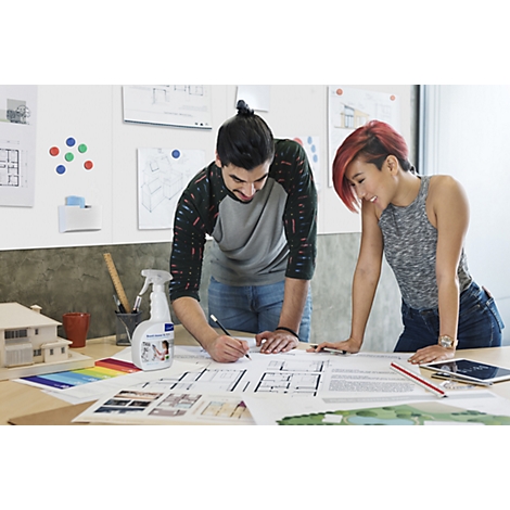 Two people are looking at construction plans. One is drawing, the other is watching. A spray bottle and color samples are on the table.