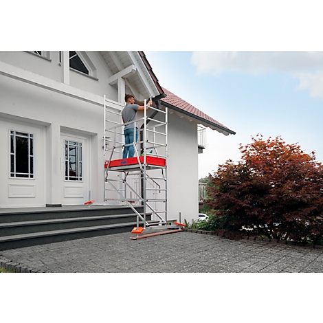 Man working on scaffolding on a house. White facade, grey paving stones, red scaffold board.