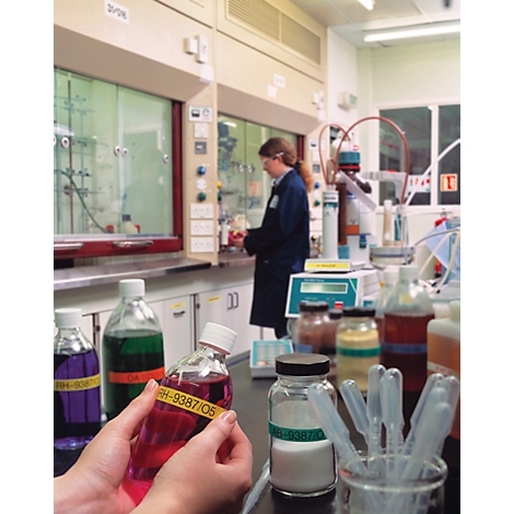 Person holding a bottle with red liquid in a lab. Other bottles with liquids are on a table, another person is working in the background.