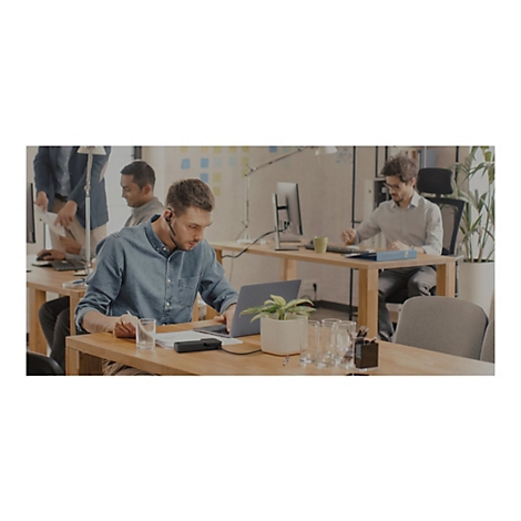 Mann mit Headset arbeitet am Laptop im Büro, Kollegen im Hintergrund. Auf dem Tisch ein Glas, eine Pflanze und ein Gerät.