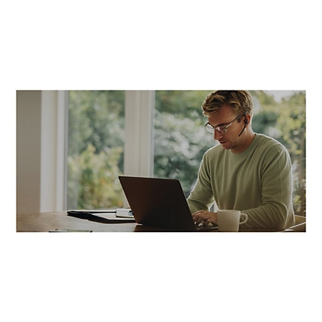 Ein Mann mit Brille und Headset arbeitet am Laptop. Auf dem Tisch liegen Unterlagen, ein Becher Kaffee steht daneben. Im Hintergrund verschwommene Bäume.