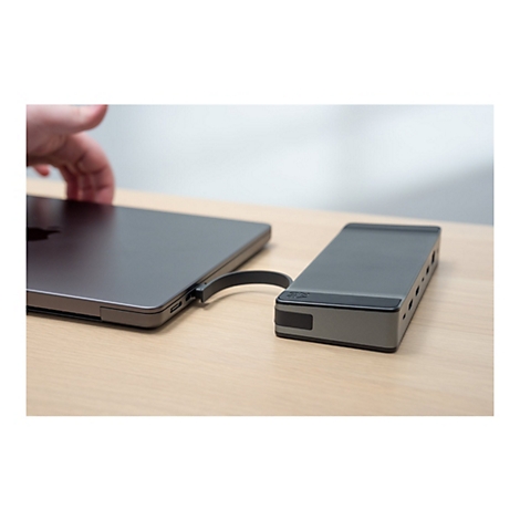 Un ordinateur portable gris et un hub gris sont reliés par un câble, sur une table en bois clair. Une main s'approche de l'ordinateur portable.