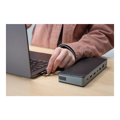 Une personne connecte un connecteur USB-C à un ordinateur portable. Une station d'accueil noire se trouve à droite. Le fond est une table en bois.