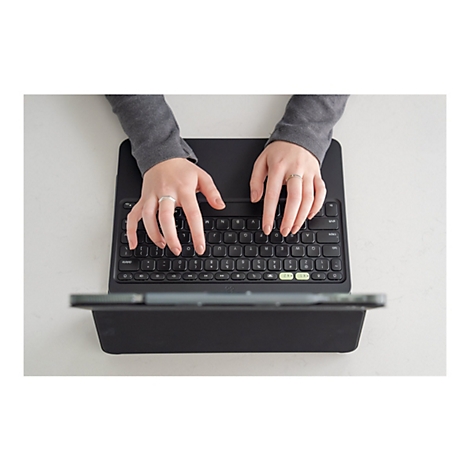 Nahaufnahme von Händen, die eine schwarze Tastatur bedienen. Die Person trägt einen grauen Pullover und Ringe an den Fingern. Die Tastatur ist mit einem Tablet verbunden und liegt auf einer weißen Oberfläche.
