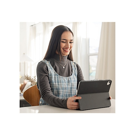 Lächelnde junge Frau, die auf einem Tablet sitzt. Sie trägt einen grauen Rollkragenpullover und eine blaue Latzhose. Auf dem Tisch befindet sich eine Vase mit dekorativen Ästen.