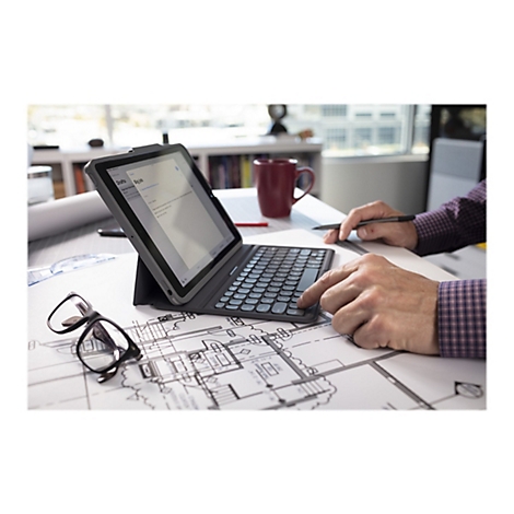 Ein Mann arbeitet an einem Tablet mit Tastatur, das auf einem Tisch mit Bauplänen liegt. Auf dem Bildschirm ist eine E-Mail-Übersicht zu sehen.