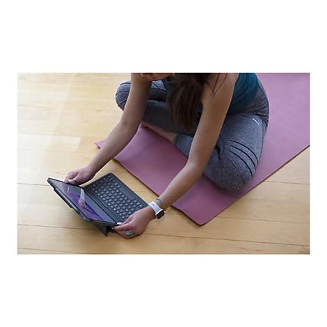 Eine Person in Yoga-Kleidung sitzt mit verschränkten Beinen auf einer Yogamatte und bedient ein Tablet mit Tastatur.