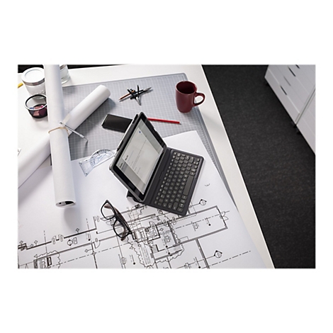 Ein Tablet mit Tastatur liegt auf einem Schreibtisch mit Bauplänen, daneben Brille und roter Becher.