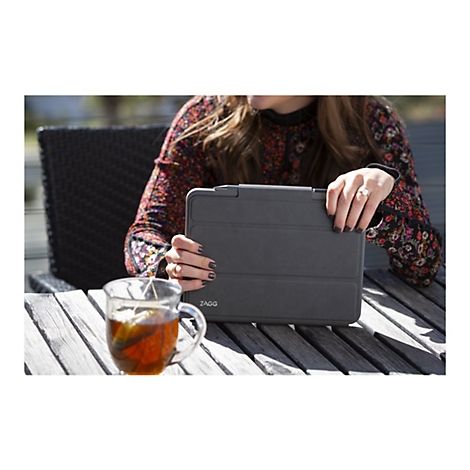 Une personne tenant un étui de tablette gris, à l'extérieur, à côté d'une tasse de thé.