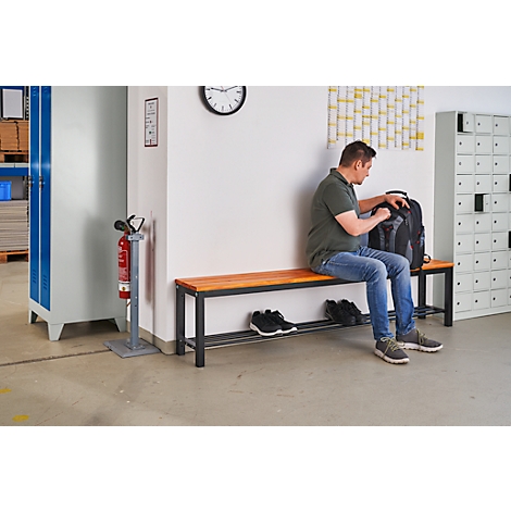 Een man zit op een bank en pakt zijn rugzak uit. Schoenen liggen op de onderste plank van de bank.
