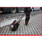 Person zieht eine schwarze Trolley-Box mit Rädern über ein Pflaster. Im Hintergrund ein Bus und ein Auto.
