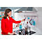 A woman in a red top in a kitchen takes a ladle from a hanging rack. A pot is on the stove.