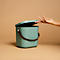 Light green trash can with open lid. A hand approaches the lid. Brown leather straps on the bucket.