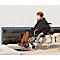 An elderly woman in a wheelchair drives over a ramp. She is wearing a black coat. In the background a window and stones.