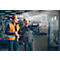 Two men in helmets and work clothes in a factory. One is pointing at a machine, the other is holding plans.