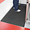 Person standing on a black perforated mat with round holes. The floor is red and gray. Feet in white shoes.