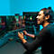 Ein junger Mann mit Dutt spielt lächelnd ein Videospiel an einem Gaming-PC mit zwei Monitoren und RGB-Beleuchtung.