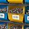 Gelbe und blaue Behälter mit Schrauben und Muttern in einem Lager. Beschriftungen mit Produktbezeichnungen und Barcodes.