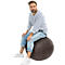 Un hombre sentado en un balón de ejercicio redondo y marrón. Lleva vaqueros, una camisa a rayas y zapatillas blancas.