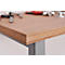 Primer plano de una mesa de madera con capas visibles, sobre una pata de mesa gris. Las herramientas están borrosas en el fondo.