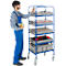 A man in blue overalls reaching into a box on a blue trolley. Grey plastic crates with tools and spare parts.