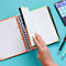 Gros plan : une personne feuillette un cahier avec des accents verts et oranges. Un téléphone et des stylos sont sur la table.