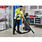 A man in a yellow jacket with a black suction system. He is vacuuming a floor in a workshop.