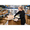 Une femme en tenue de travail ouvre avec concentration un carton d'expédition dans un entrepôt entre des rayonnages à palettes.