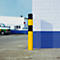 Yellow and black bollard in front of white and blue brickwork. Vehicles and a gate in the background.