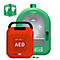 Roter AED-Defibrillator mit Tragegriff neben einem grünen Gehäuse. Auf dem AED steht in Weiß „AED“. Oben rechts ein Schild mit Anweisungen.