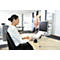 A woman sits at a desk, typing on a keyboard, and looking at a monitor. On the desk is a monitor stand with documents.