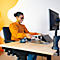 A smiling woman in an orange blouse sits at a height-adjustable desk, holding papers and looking at a laptop.