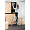 Une femme sur un escabeau sort des dossiers d'une armoire. Elle porte une blouse claire et un pantalon noir.