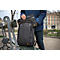 Mann hält Rucksack und Laptoptasche. Beide schwarz mit gelben Akzenten und Logo. Im Hintergrund Park mit Bank.
