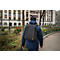 Ein Mann mit schwarzem Rucksack geht einen Weg entlang, Gebäude im Hintergrund.
