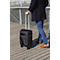 Person zieht einen schwarzen Trolley auf Holzstegen. Er trägt einen dunklen Mantel, blaue Hose und braune Schuhe.