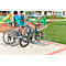 Drei Fahrräder stehen auf einem Gehweg in einem Fahrradständer. Im Hintergrund befindet sich ein Spielplatz.