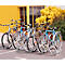 Mehrere Fahrräder stehen in einem Fahrradständer im Freien. Im Hintergrund ein gelbes Gebäude und Grünflächen.