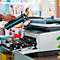 Eine Hand hält eine blaue Tonerkartusche über einem Drucker. Eine Person ist im Hintergrund unscharf zu sehen.