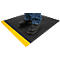 A person stands on a black anti-slip mat with a yellow edge. Feet are in black shoes and jeans.