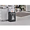 A black paper shredder stands next to a desk. Paper is being shredded at the top, and the container below is filled with shredded paper.