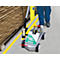 A person uses a road marking machine to draw a yellow line on the floor. In the background, a shelf and paint sprays can be seen.