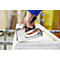 A hand holding a silver staple gun, pointed at a white wooden frame.