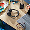 Black headset on a wooden desk, next to a laptop and glasses. In the background notes and a charging station.