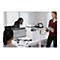Büro mit drei Personen. Ein Drucker steht auf einem Schreibtisch, eine Frau hält Papier, eine andere schaut auf einen Monitor.