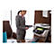 Eine Frau in einem grauen Anzug steht an einem Schreibtisch und hält ein Tablet vor einem Drucker. Der Drucker, eine HP OfficeJet Pro 7740, ist geöffnet und druckt ein Dokument.