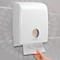 Dispensador de toallas de papel blanco en una pared gris. Una mano saca una toalla de papel.